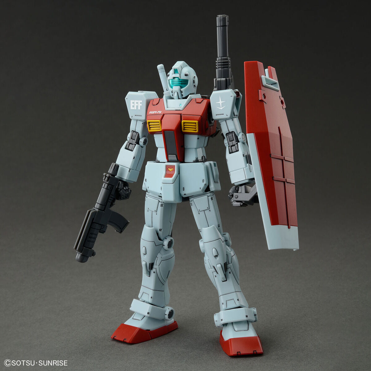 Model kit Bandai HG 1/144 GM con cannone a spalla, fucile e scudo, in posa frontale su sfondo grigio.