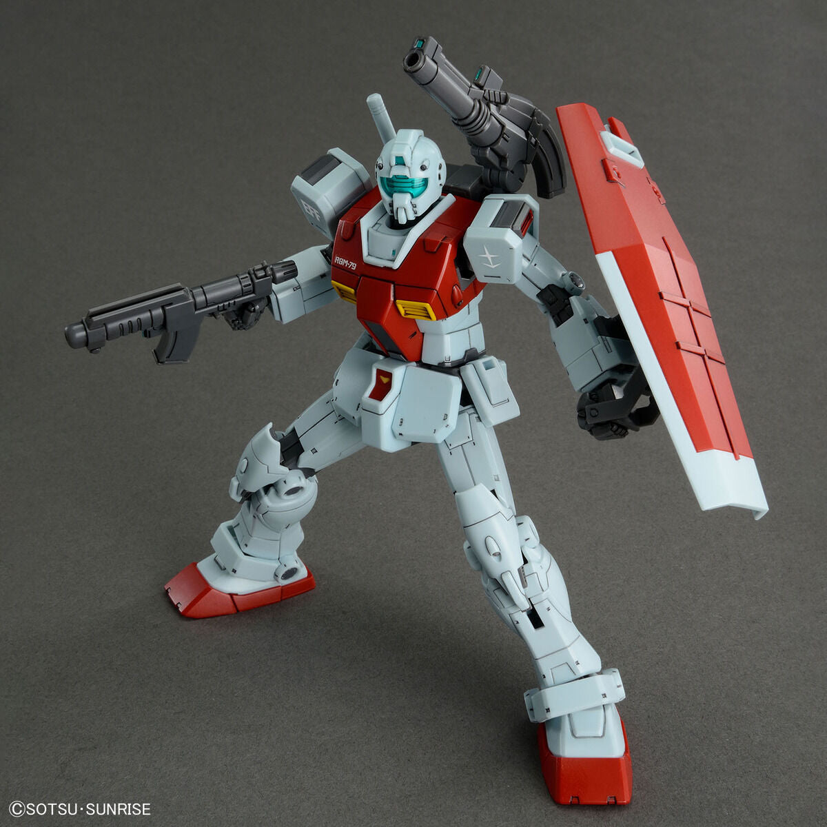 Model kit Bandai HG 1/144 GM con cannone a spalla, scudo e fucile, in posa dinamica su sfondo grigio.